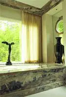 Bathroom with a large marble-clad tub, sheer yellow curtains, and a dark vase on a windowsill overlooking green trees.