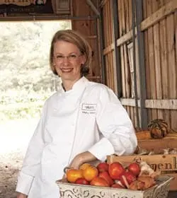 Woman in chef's coat standing in a rustic barn with a basket of tomatoes and gourds.
