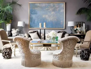 Living room with plush seating, a large abstract blue painting, and lush greenery. Two ornate chairs with fringe face a sofa and glass coffee table.