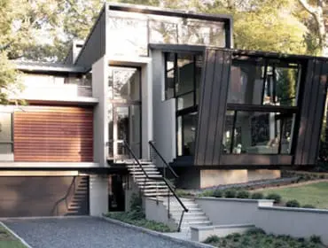 Modern house with geometric design, featuring large windows, wood siding, metal panels, and a prominent staircase leading to the entrance.