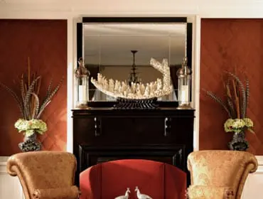 Living room with two ornate gold armchairs flanking a red sofa, a dark wood fireplace with a mirrored surround, and a glass-top coffee table featuring two peacock figurines.