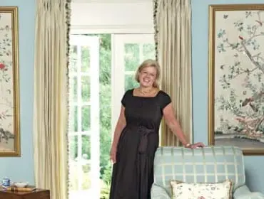 Woman in a dark dress stands in a room with light blue walls, leaning against a light blue plaid armchair. French doors open to a garden.