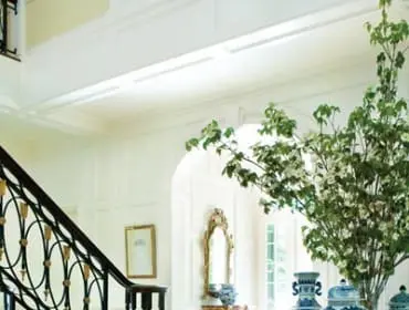 Foyer with a grand staircase featuring ornate black and gold railings, a cream upholstered chair with a blue and green patterned pillow, and a round table draped in a fringed yellow damask cloth adorned with blue and white porcelain vases.