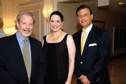 Three people posing for a photo indoors. The person on the left has a beard and is wearing a suit and tie. The person in the middle is a woman wearing a black dress and a gold necklace. The person on the right is a man wearing a suit and tie.