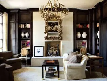 Living room with dark wood built-in bookshelves, a stone fireplace, ornate gold mirror, and plush seating including a cream sofa and brown armchairs.