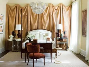 Bedroom with a four-poster bed, draped gold fabric backdrop, ornate chandelier, and a velvet armchair in the foreground.