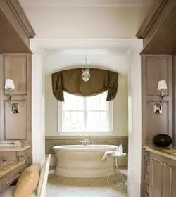 Neutral bathroom with a freestanding tub under a window with a valance, flanked by wood cabinetry and a vanity area with a chair.
