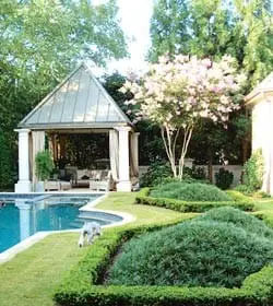 Curving swimming pool with a covered cabana and manicured hedges surrounding lush green landscaping and a flowering tree.