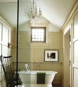 Cream paneled bathroom with a freestanding clawfoot tub, glass shower enclosure, black Windsor chair, and crystal chandelier.