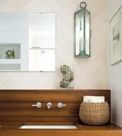 Modern bathroom vanity with a wooden countertop and integrated sink, brushed nickel fixtures, and a woven basket holding towels. A lantern light hangs on a herringbone tile wall.