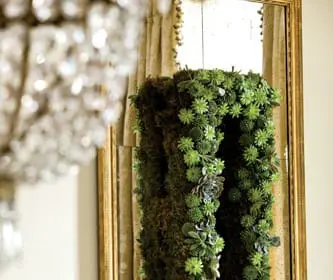 A vertical living wall art piece made of moss and succulents hangs in front of a tall, ornate gold-framed mirror with gold damask curtains behind it.