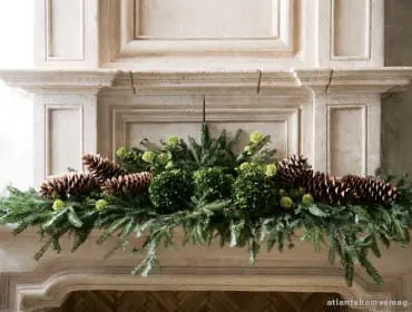Stone fireplace mantel decorated with a festive garland of pine branches, pinecones, boxwood spheres, and green hydrangeas.
