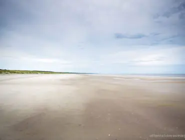Wide sandy beach with dunes covered in green grass and the ocean under a cloudy sky.