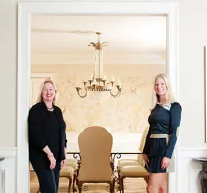 Two women stand in a doorway. The woman on the left wears a black suit. The woman on the right wears a dark blue dress. Behind them is a dining room with a chandelier and floral wallpaper.