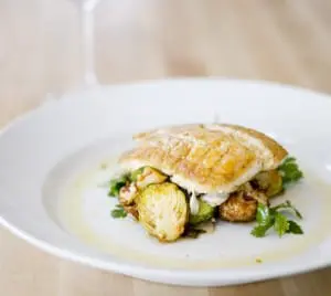 Close-up of a white plate with a golden-brown pastry topped with roasted Brussels sprouts and herbs, drizzled with oil.
