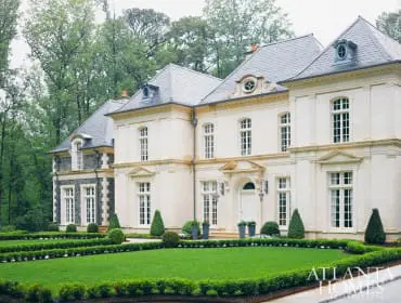 French-inspired mansion with cream stone facade, gray slate roof, manicured hedges, and a lush green lawn.