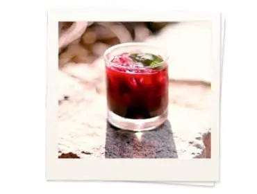 Close-up of a deep red cocktail in a clear glass with ice and a green garnish, resting on a textured stone surface.