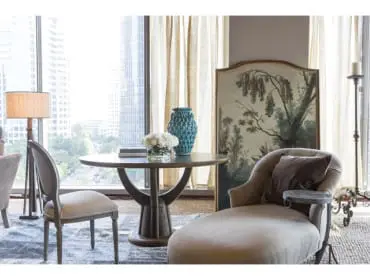 Living room with a round wooden table, two chairs, a chaise lounge, and a floor lamp, with a large window overlooking a city skyline.