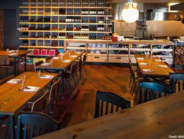 Restaurant dining room with wooden tables and chairs, extensive wine shelving, and an open kitchen area in the background.