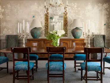 Dining room with a long wooden table and chairs upholstered in blue fabric. The walls feature a neutral wallpaper with a tree and bird motif, and a large ornate mirror hangs above a wooden sideboard.
