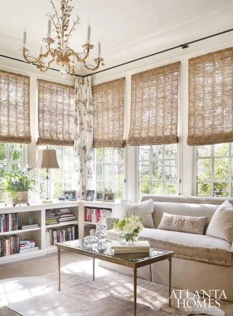 Light-filled living room with built-in bookshelves, a neutral sofa with decorative pillows, a glass-top coffee table, and woven Roman shades covering large windows.