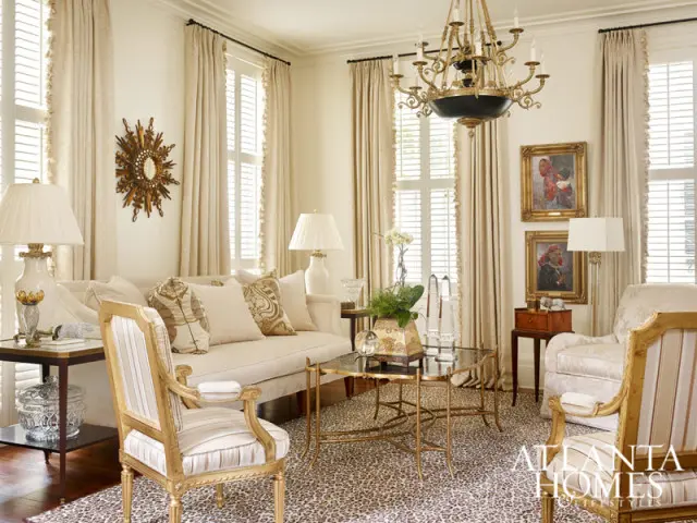 Living room with cream sofa, patterned pillows, gold accents, leopard print rug, and ornate chandelier. Two framed portraits hang on the wall.