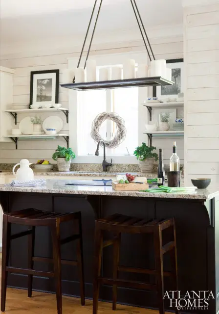 Dark wood kitchen island with granite countertop, two wooden bar stools, and a sink with a wreath above it. White shiplap walls and open shelving with dishes are visible.