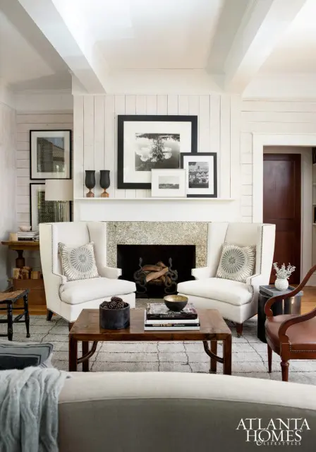 Living room with white wingback chairs flanking a stone fireplace, a dark wood coffee table, and framed black and white photographs on the wall.