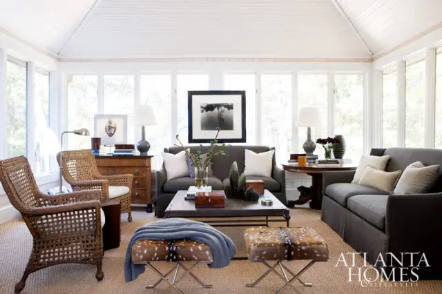Sunroom with vaulted white ceiling, large windows, charcoal sofas, wicker chairs, a dark wood coffee table, and two animal print ottomans.