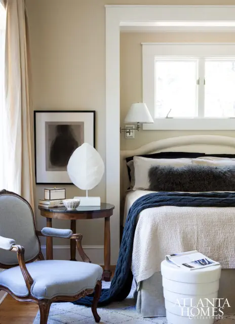 Bedroom with a light beige upholstered headboard, white bedding, a dark blue cable-knit throw, and a gray fur lumbar pillow. A vintage armchair sits beside a round side table with a framed artwork and a white sculpture.