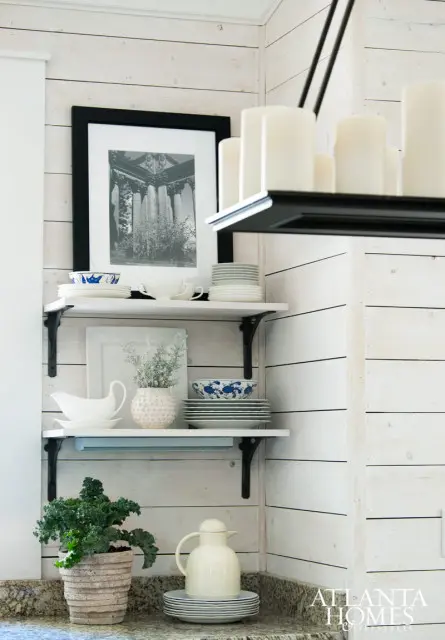 White shiplap walls with open shelving displaying white plates, blue and white patterned bowls, a white gravy boat, a textured vase with dried flowers, and a framed black and white print of columns.