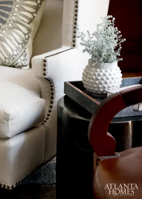 Upholstered armchair in light beige fabric with nailhead trim, a decorative pillow with a gray and white pattern, and a textured white vase with a plant on a dark tray.