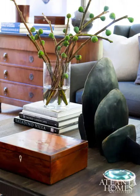 Wooden coffee table with a decorative box, stack of books, glass vase with fig branches, and dark green abstract sculptures. Atlanta Homes & Lifestyles logo in the corner.