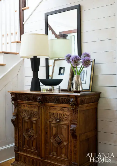 Ornate wooden cabinet with carved details topped with a black lamp, framed art, a silver vase with purple flowers, and a black bowl on a clear stand.