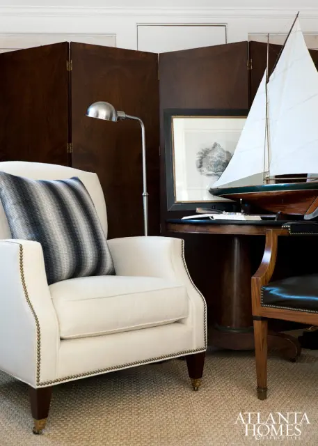 Cream upholstered armchair with striped pillow, dark wood folding screen, model sailboat, and framed artwork on a round table.