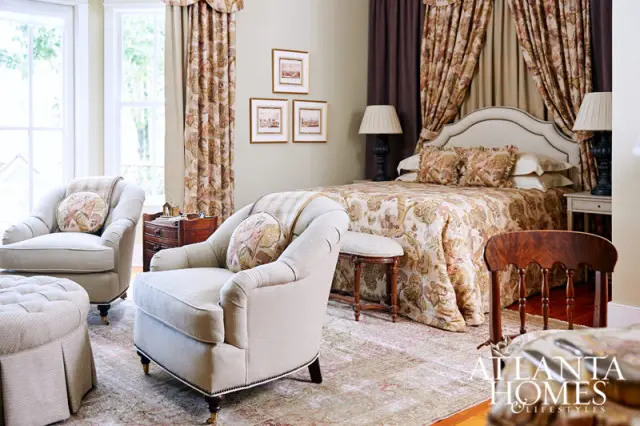 Bedroom with a large bed dressed in floral print comforter and pillows, flanked by two lamps. Two upholstered armchairs and an ottoman sit in the foreground.