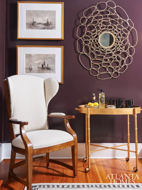 Wingback chair with cream upholstery next to a gold bar cart with drinks and lemons, beneath two framed nautical prints and a decorative mirror on a plum wall.