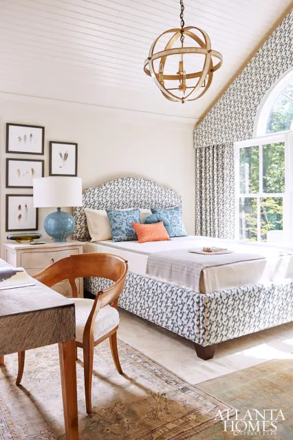 Bedroom with a patterned upholstered bed and headboard, blue and orange accent pillows, a wooden desk and chair, and a large arched window with patterned curtains.