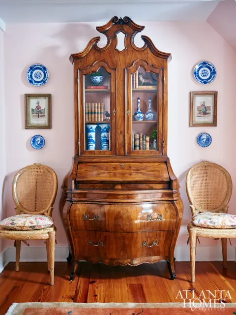 Antique wooden secretary desk with glass doors flanked by two cane-backed chairs with floral cushions against a pale pink wall. Blue and white porcelain plates and framed art adorn the wall.