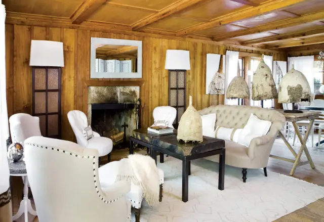 Living room with wood-paneled walls and ceiling, stone fireplace, tufted sofa, white armchairs, and a dark metal coffee table with decorative objects.