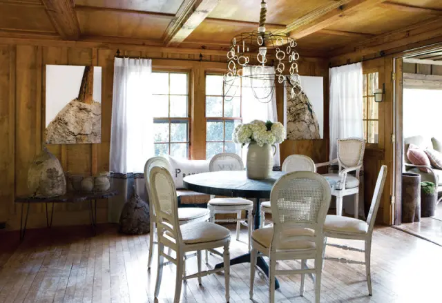 Round dining table with cane-back chairs in a wood-paneled room with beamed ceiling and abstract art.