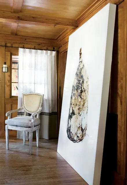 Wooden paneled room with a large abstract artwork leaning against the wall, a white upholstered chair with nailhead trim, and sheer curtains covering a window.