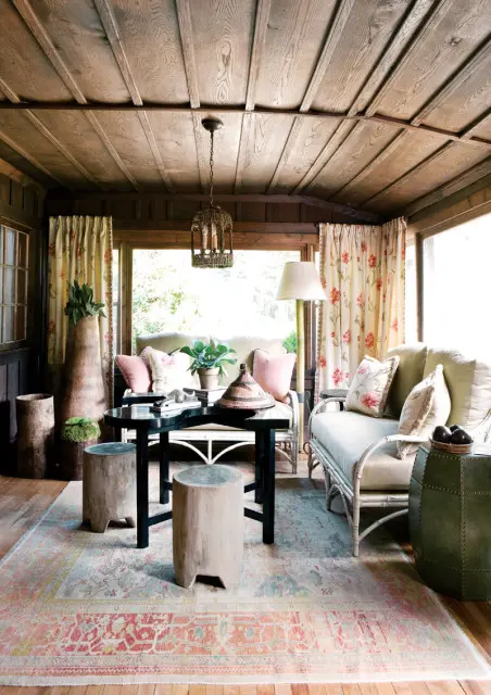 Sunroom with wood-paneled walls and ceiling, featuring a black coffee table, two white wicker sofas, floral curtains, and a patterned rug.