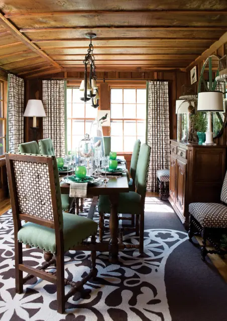 Dining room with wood-paneled walls and ceiling, featuring a dark wood table set with green glassware and white patterned curtains.