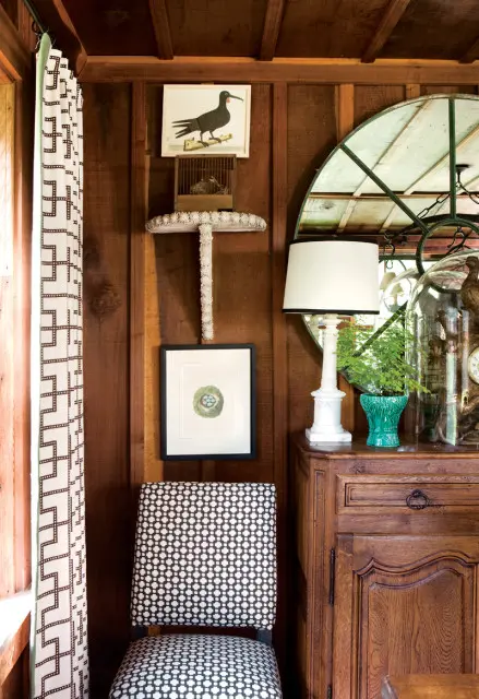 Wooden paneled room with a black and white patterned chair, botanical prints of a bird and nest, and a white lamp on an antique wooden cabinet.