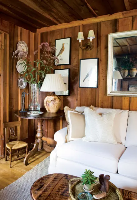 Living room with wood-paneled walls, a white sofa with neutral pillows, and a round wooden side table with a lamp and floral arrangement.