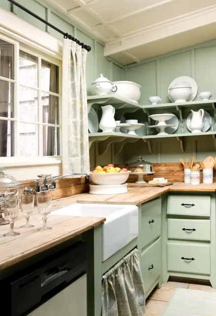 Sage green kitchen with white farmhouse sink, butcher block countertops, open shelving displaying white dishware, and a window with patterned curtains.