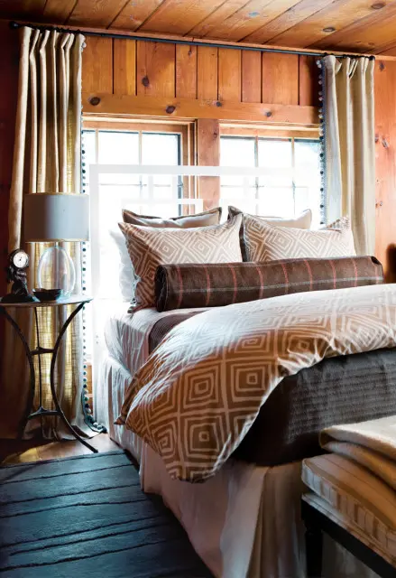 Bedroom with wood-paneled walls and ceiling, featuring a white bed with geometric patterned bedding, bolster pillow, and decorative curtains.