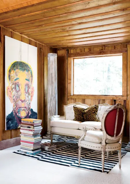 Living room with wood-paneled walls and ceiling, featuring a large abstract portrait, a white tufted sofa with a leopard print pillow, a distressed armchair with red upholstery, and a black and white striped rug.