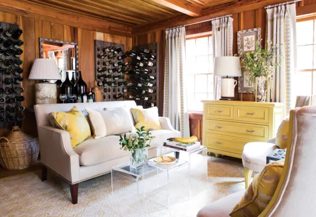 Living room with wood-paneled walls, cream sofa with yellow and monogrammed pillows, and a yellow dresser. Wine racks line the walls.